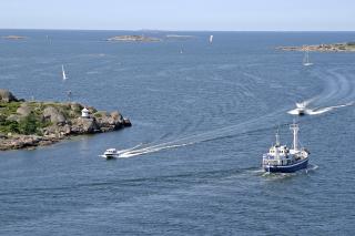 Kesäinen saaristokuva, jossa merta, luotoa sekä laiva ja kaksi venettä.